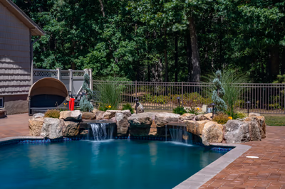 Multi-tiered natural stone boulder waterfalls cascading into a swimming pool surrounded by lush greenery