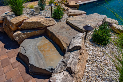Multi-tiered natural stone boulder waterfall cascading into a pool surrounded by lush garden plants