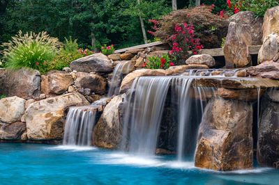 Multi-tiered natural stone boulder waterfalls cascade into a pool surrounded by lush, vibrant landscaping