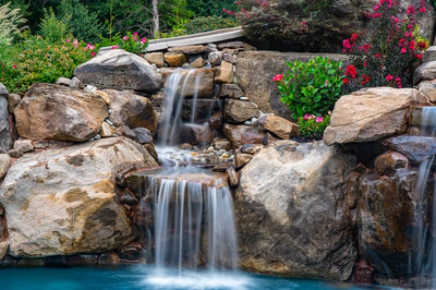 Multi-tiered natural stone boulder waterfall cascading into a pool surrounded by lush garden plants