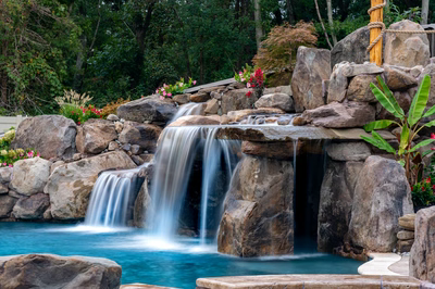 Multi-tiered natural stone boulder waterfalls cascade into a pool surrounded by lush garden plants