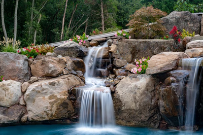 Multi-tiered natural stone boulder waterfall cascading into a pool surrounded by lush flowering plants