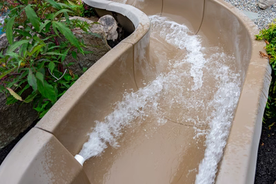 Multi-tiered natural stone boulder waterfall with rushing water streams beside lush plants and pool
