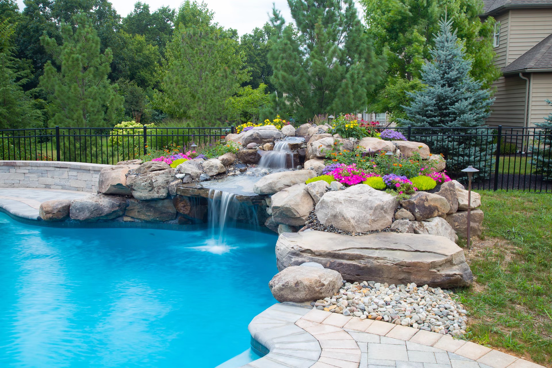 Two-tiered natural stone boulder waterfall cascading into a pool surrounded by colorful garden plants