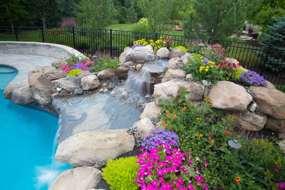 Two-tiered natural stone boulder waterfall cascades into a pool surrounded by vibrant garden flowers