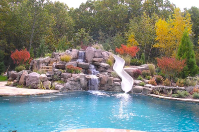 Three-tiered natural stone boulder waterfall cascading into a pool amidst colorful trees and lush landscaping