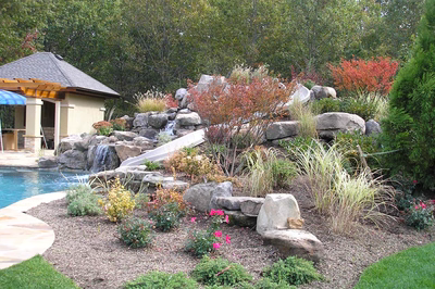 Multi-tiered natural stone boulder waterfall cascading into a pool surrounded by lush garden landscaping