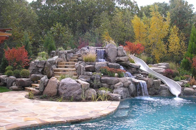 Multi-tiered natural stone boulder waterfall cascading into a pool surrounded by lush, colorful landscaping