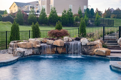 Twin natural stone boulder waterfalls cascade into a backyard pool surrounded by lush green shrubs