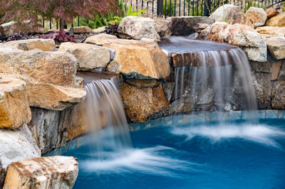 Two-tiered natural stone boulder waterfall cascading into a pool surrounded by lush garden plants