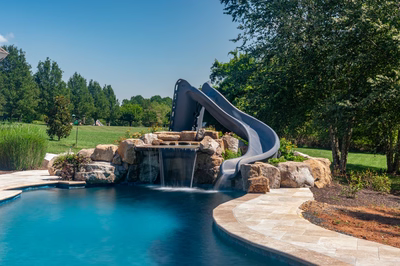 Natural stone boulder waterfall cascading in two tiers into a pool beside slide and trees