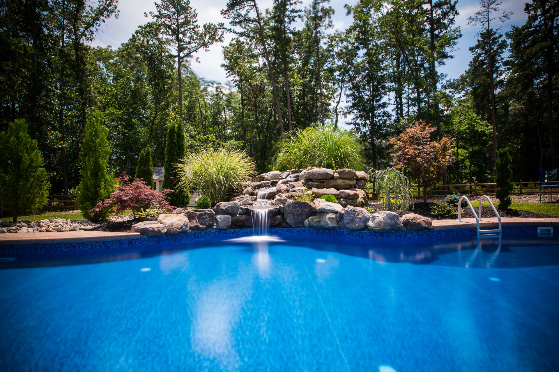 Multi-tiered natural stone boulder waterfall cascading into a blue pool bordered by lush landscaping