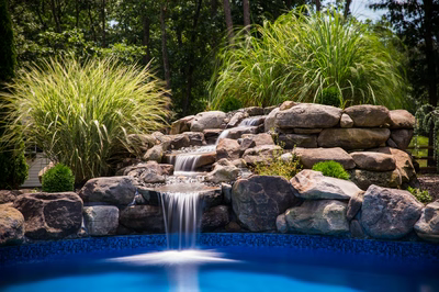 Three-tiered natural stone boulder waterfall cascading into a blue pool with lush ornamental grasses