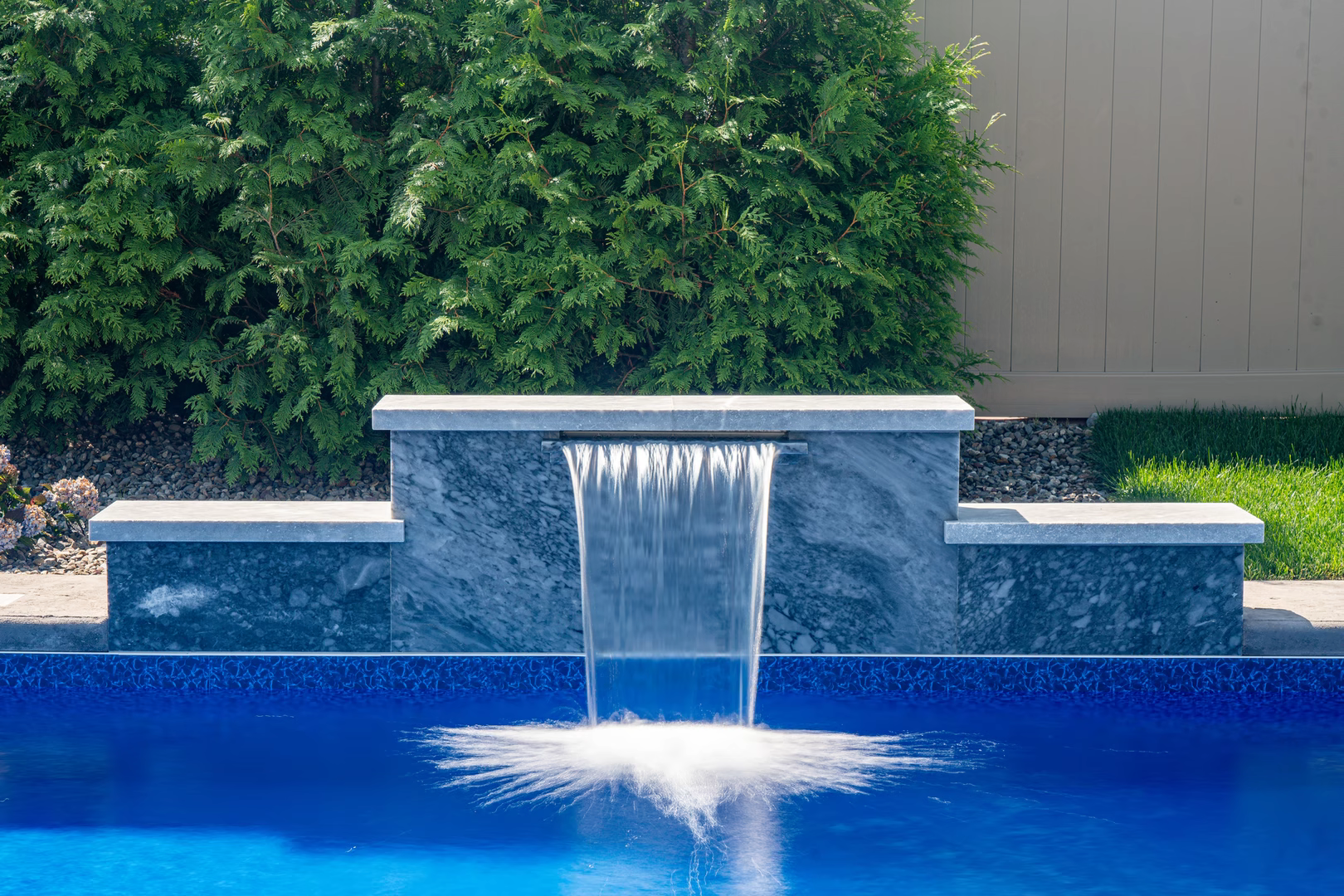 Natural stone wall featuring sheer descent waterfall spilling into pool with lush hedges
