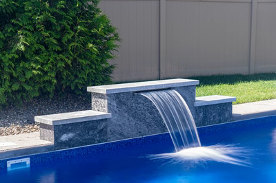 Three-tiered natural stone boulder waterfall cascading into a swimming pool beside a green shrub