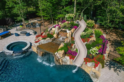 Multi-tiered natural stone boulder waterfall cascading into a swimming pool surrounded by vibrant floral landscaping