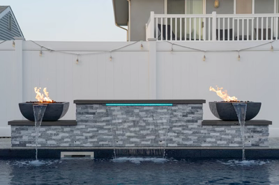 Three-tiered natural stone waterfalls with fire bowls cascading into a pool against white fencing