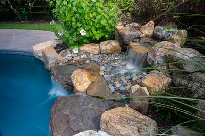 Multi-tiered natural stone boulder waterfall cascading into a pool surrounded by lush garden greenery