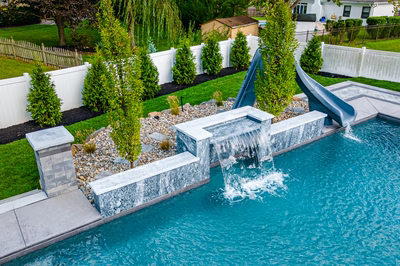 Single-tier sheer descent stone waterfall cascading into a backyard pool surrounded by lush evergreen trees