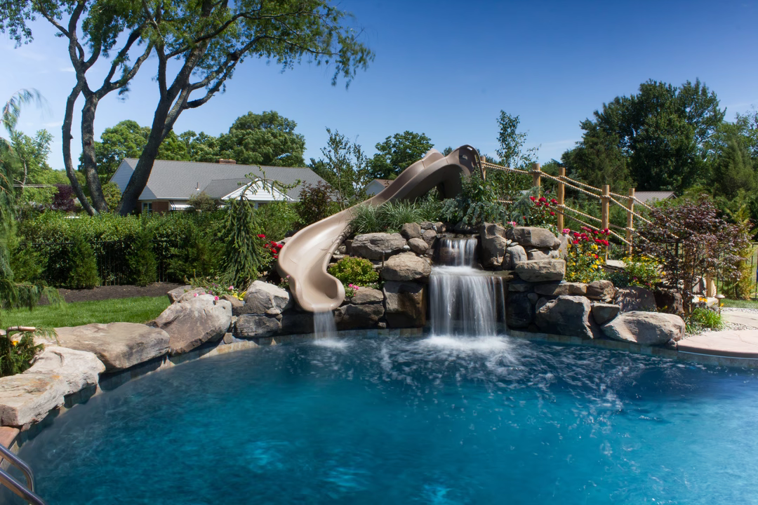 Two-tiered natural stone boulder waterfall cascading into a swimming pool surrounded by lush garden landscaping