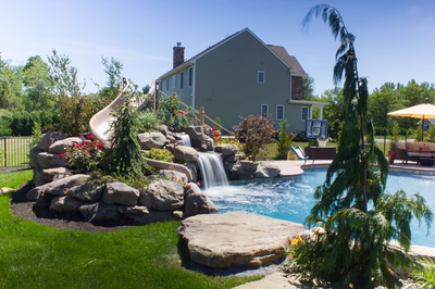 Multi-tiered natural stone boulder waterfall cascading into a swimming pool surrounded by lush landscaping