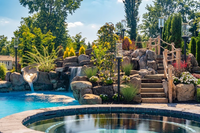 Two-tiered natural stone boulder waterfall cascading into a pool surrounded by lush, vibrant garden plants