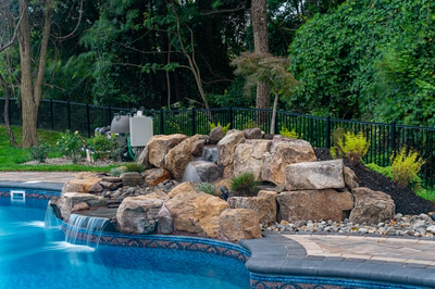 Multi-tiered natural stone boulder waterfall cascading into a backyard pool amidst lush garden plants