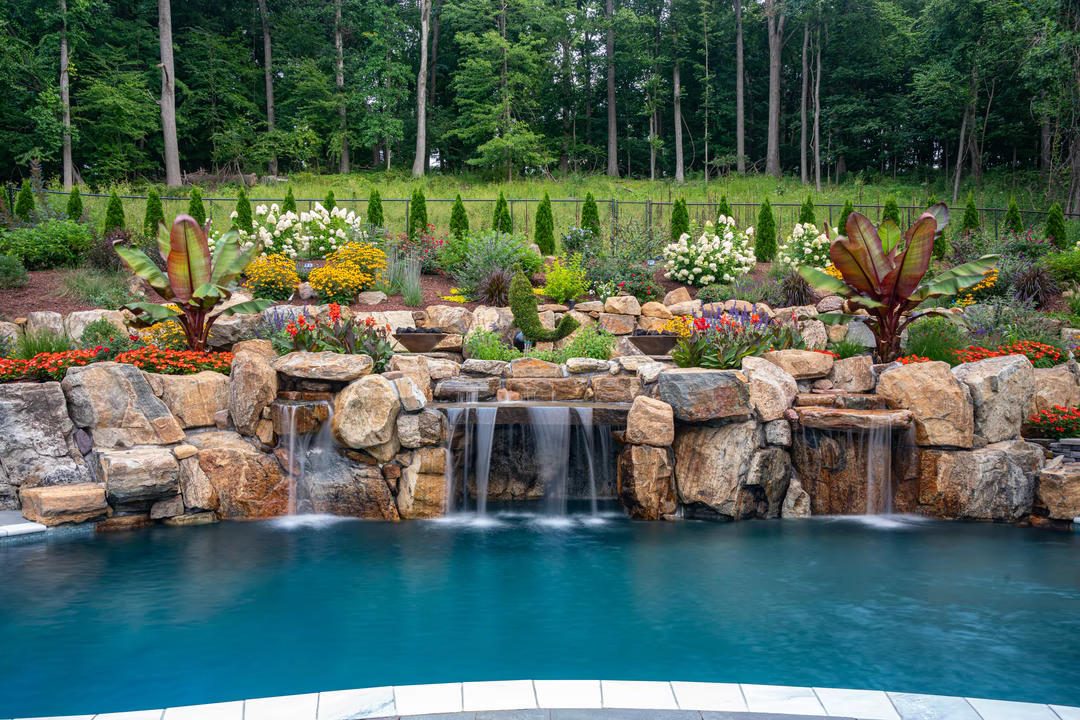 Multi-tiered natural stone boulder waterfalls cascading into a pool surrounded by lush, colorful landscaping