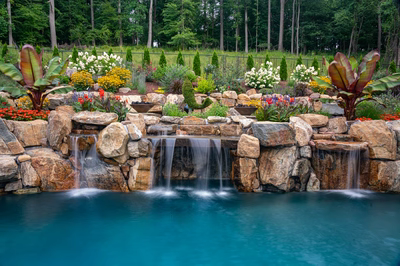 Multi-tiered natural stone boulder waterfalls cascading into a pool surrounded by lush, vibrant landscaping