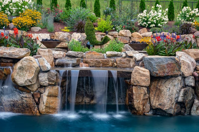 Two-tiered natural stone boulder waterfall cascading into a pool surrounded by lush garden landscaping