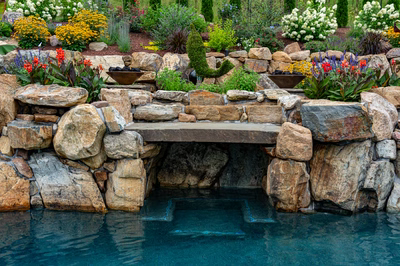 Multi-tiered natural stone boulder waterfall cascading into a pool surrounded by vibrant garden plants
