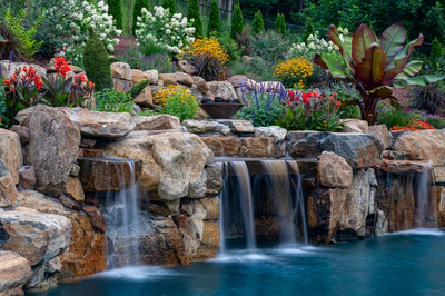 Triple-tiered natural stone boulder waterfalls cascading into a pool amidst lush garden landscaping