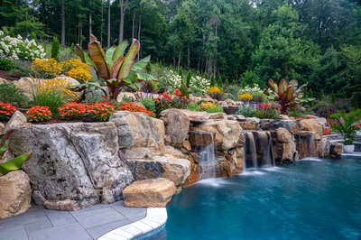 Multi-tiered natural stone waterfalls cascade into a pool surrounded by lush, colorful tropical landscaping