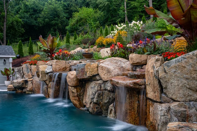Multi-tiered natural stone boulder waterfalls cascading into a pool surrounded by lush, colorful garden landscaping