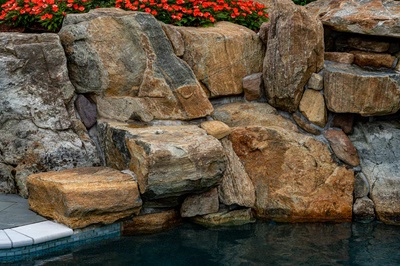 Multi-tiered natural stone boulder waterfall cascading into a pool with vibrant red flowers