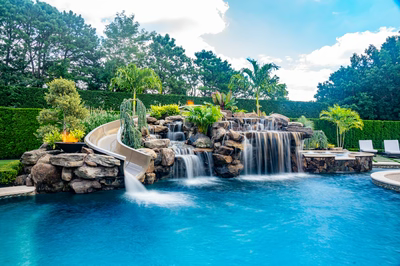 Multi-tiered natural stone boulder waterfall cascading into a pool with tropical plants and slide