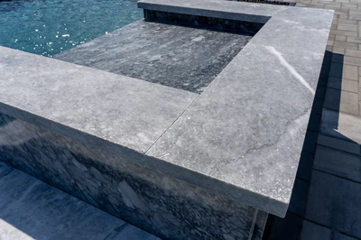 Draft 5: Single-tiered grey marble waterfall cascading into a clear swimming pool beside a paved patio.
*   Word count: 1. Single-tiered 2. grey