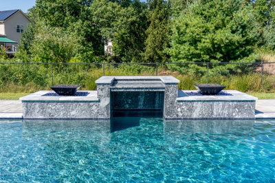 Modern three-tiered stone waterfall feature spilling into a swimming pool with lush green foliage