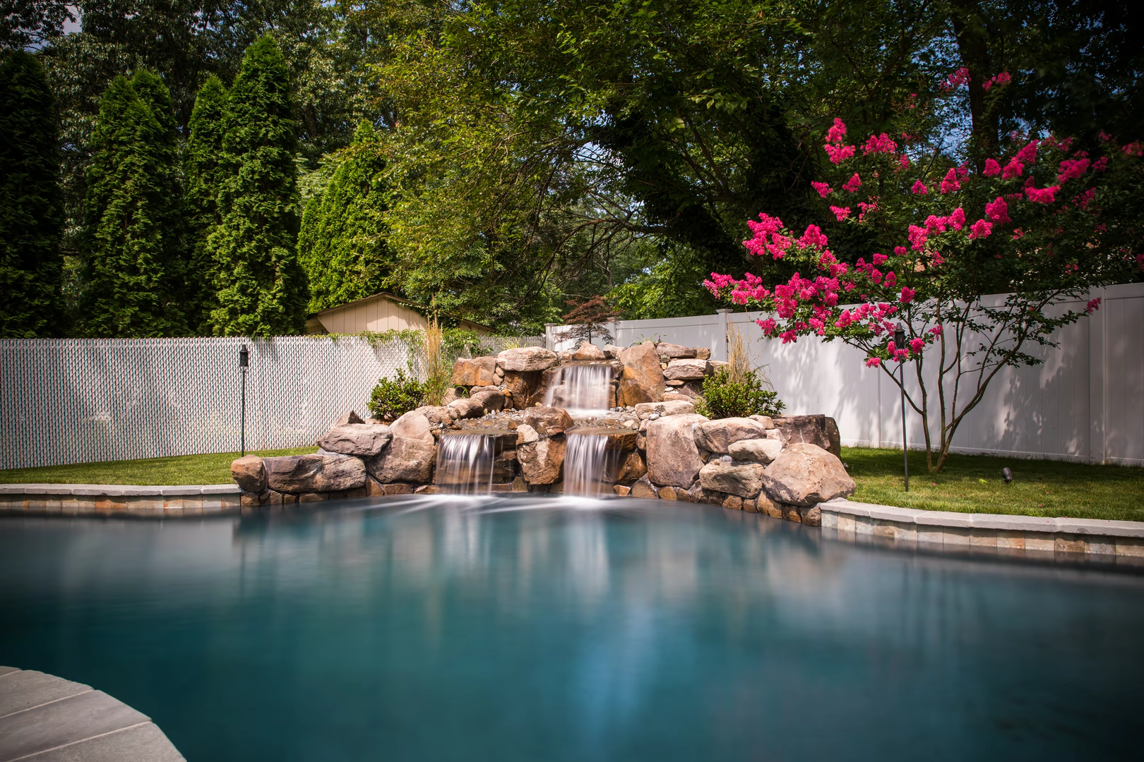 Natural stone boulder waterfall cascading into a pool beside vibrant pink flowering trees