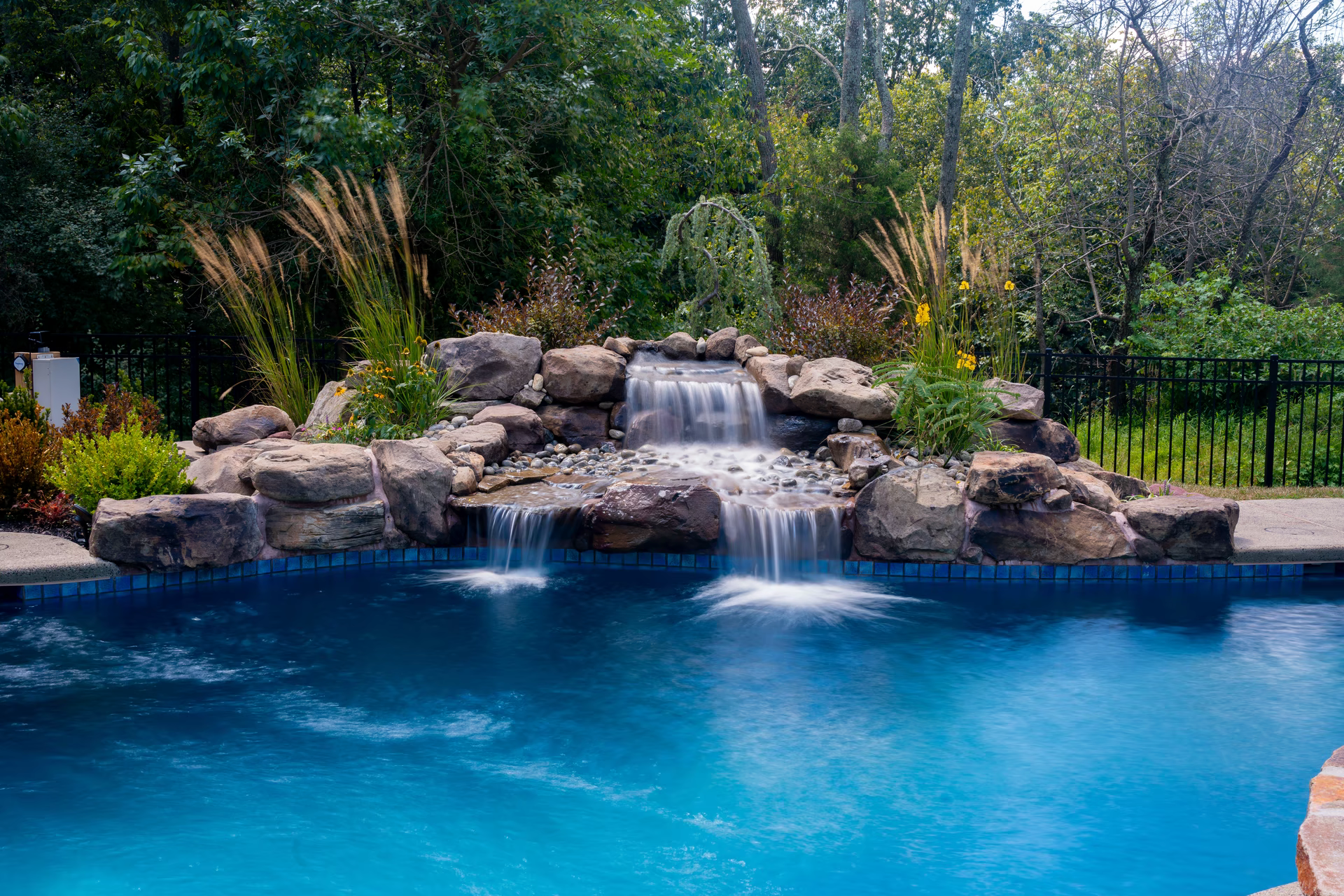 Multi-tiered natural stone boulder waterfall cascading into a pool surrounded by lush garden greenery