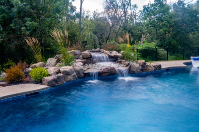 Two-tiered natural stone boulder waterfall cascading into a pool surrounded by lush ornamental greenery