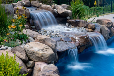 Multi-tiered natural stone boulder waterfall cascades into a swimming pool amidst lush garden plants