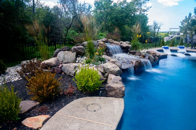 Two-tiered natural stone boulder waterfall cascading into a swimming pool with lush garden plants