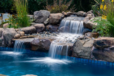 Multi-tiered natural stone boulder waterfall cascading into a pool surrounded by lush garden plants
