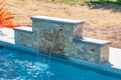 Three-tiered natural stone waterfall cascading into a swimming pool with dirt and grass surroundings