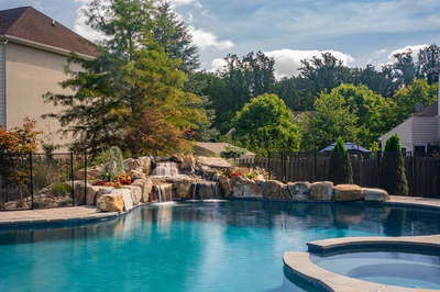 Multi-tiered natural stone boulder waterfalls cascading into a backyard pool surrounded by lush green trees