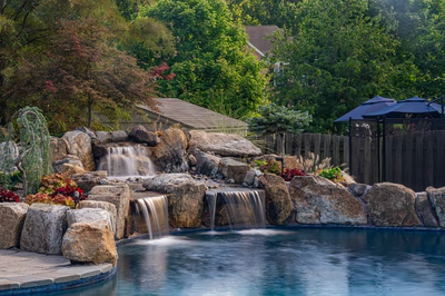 Two-tiered natural stone boulder waterfall cascading into a pool surrounded by lush garden greenery