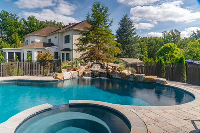 Multi-tiered natural stone boulder waterfall cascading into a backyard pool surrounded by lush greenery