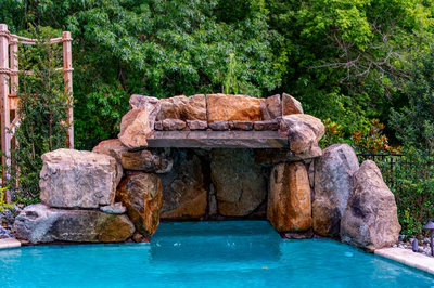 Multi-tiered natural stone boulder waterfall cascading into a pool surrounded by dense green landscaping