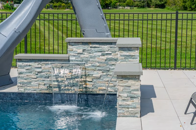 Multi-tiered natural stone waterfall cascading into a backyard pool near a lawn and fence